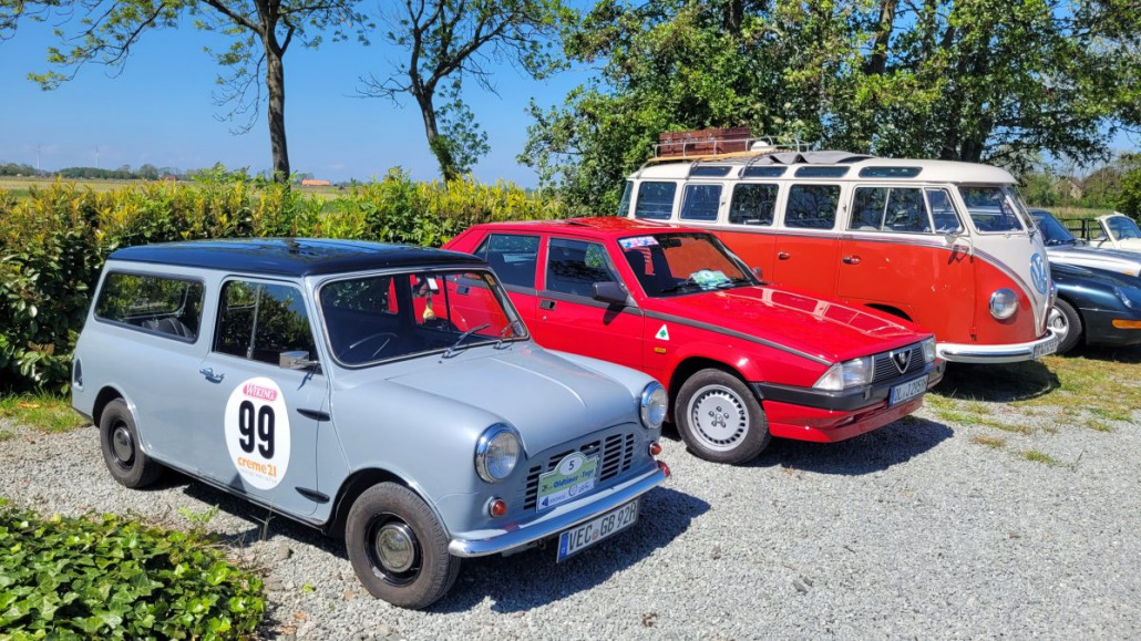 Ein grauer klassischer Mini Cooper, ein roter Alfa Romeo-Schrägheckwagen und ein orangefarbener Volkswagen-Bus sind auf einer Rasenfläche mit Bäumen im Hintergrund geparkt.